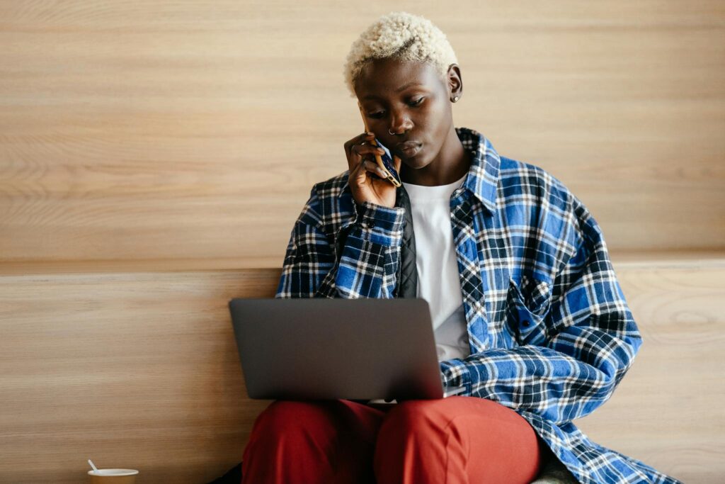 Serious African American female in trendy blue checkered shirt speaking on mobile phone while browsing netbook